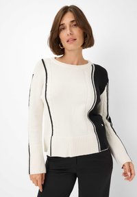 Femme portant un pull en tricot noir et blanc avec des finitions noires et des manches évasées, associé à un pantalon noir, se tenant devant un fond blanc.