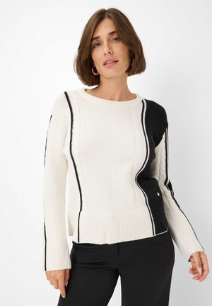 Vrouw in een wit en zwart kabelgebreid trui met zwarte randen en uitlopende mouwen, gecombineerd met zwarte broek, staande tegen een witte achtergrond.