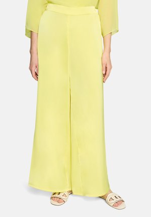 Geel wijde broekspijpen gecombineerd met crèmekleurige sandalen met open teen, gedragen door een staand persoon met ontspannen handen langs het lichaam.