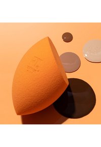 Éponge à maquillage orange à côté de quatre gouttes de fond de teint liquide dans différentes teintes de peau sur un fond orange assorti.