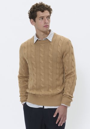 Beige kabelstickad tröja med rund halsringning, med ribbade ärmslut och nederkant, lager på en vit och blå randig skjorta.