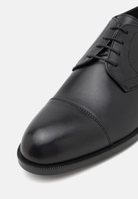 Chaussure de ville en cuir noir avec laçage fermé, bout droit et fines coutures sur la partie supérieure de la chaussure.
