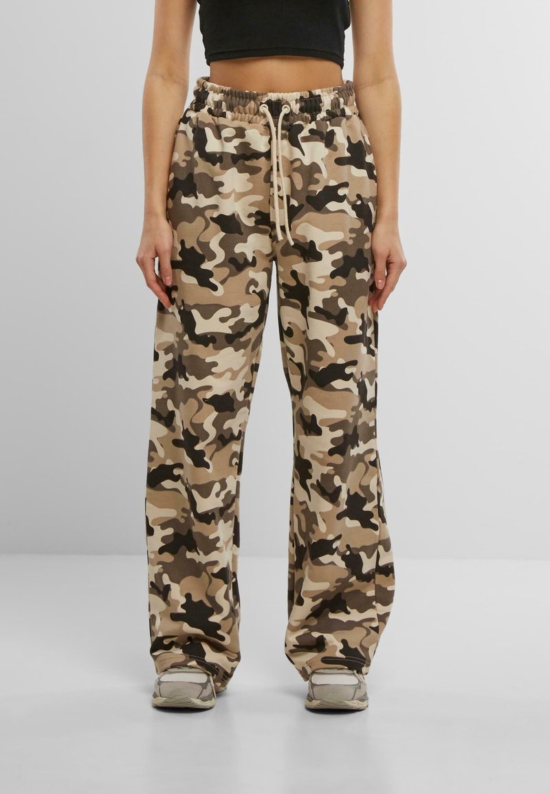 Personne portant un pantalon camouflage beige, marron et noir avec taille élastique et cordon, associé à des baskets grises et un top court noir.