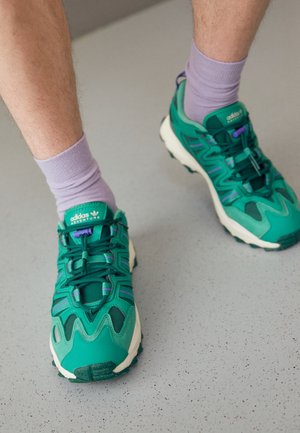 Personne portant des baskets Adidas Adventure vertes avec des accents violets et des chaussettes lilas, debout sur un sol gris moucheté.
