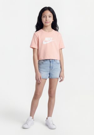 Mädchen mit langem schwarzen Haar trägt ein rosa Nike-T-Shirt, hellblaue Jeansshorts und weiße Turnschuhe, steht vor einem schlichten weißen Hintergrund.