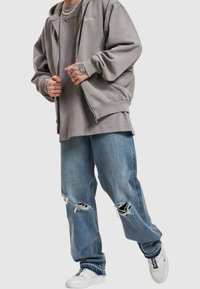 Persona vistiendo una sudadera con cremallera gris, camiseta gris de gran tamaño, jeans azules rasgados, zapatillas blancas y collares de cadena de plata, de pie contra un fondo simple.