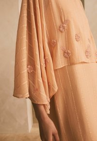 Persikofärgad klänning med strukturerade ränder, med blommig broderi i ljusrosa. Utrustad med utsvängda ärmar och en avslappnad passform som skapar ett flödande rörelse.