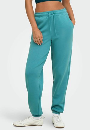Vrouw draagt turquoise joggerbroek met trekkoord in de taille, witte sneakers en een donker kort topje, staat met één hand in de zak.