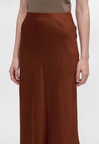 Femme portant une jupe taille haute couleur rouille et un top beige sans manches, mains détendues le long du corps sur un fond uni.