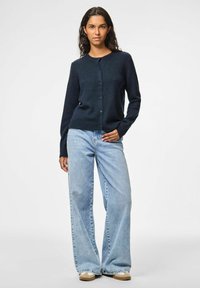 Tumši zils cardigan ar adītu tekstūru, apaļu kaklu, pogām priekšā un garām piedurknēm, apvienots ar gaiši ziliem platām kājām džinsiem.