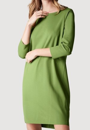 Frau trägt ein knielanges, grünes, langärmliges Kleid mit rundem Ausschnitt und posiert mit einer Hand nahe am Schlüsselbein und der anderen über der Taille.