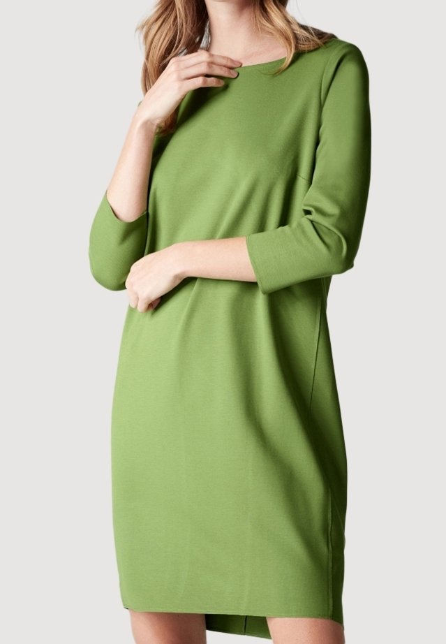 Femme portant une robe verte longue jusqu’aux genoux, à manches longues et col rond, posant une main près de sa clavicule et l’autre sur sa taille.