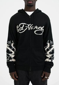 Haut de survêtement noir zippé avec le logo "Ed Hardy" brodé en blanc à l'avant, présentant des motifs complexes sur les manches, fabriqué en matériau tricoté.
