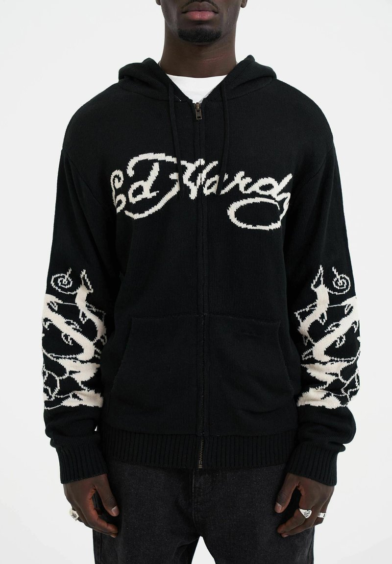 Haut de survêtement noir zippé avec le logo "Ed Hardy" brodé en blanc à l'avant, présentant des motifs complexes sur les manches, fabriqué en matériau tricoté.