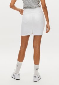 Witte denim rok met een split aan de achterkant, ontworpen in een getailleerde vorm en voorzien van riemlussen. Draagt bij witte sneakers en een gestreept grijs shirt.