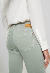 Jeans de mezclilla verde claro con bolsillos traseros y detalles cosidos, que cuentan con una etiqueta de marca de cuero marrón en la cintura y una blusa blanca.