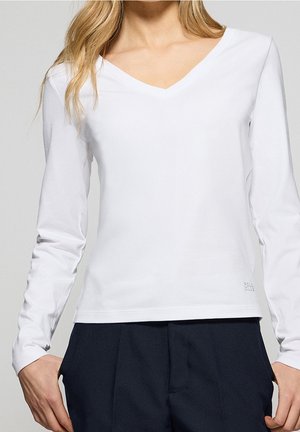 Femme portant un chemisier blanc à col en V avec manches longues et un pantalon bleu marine foncé, mains dans les poches, cheveux blonds lâchés sur une épaule.