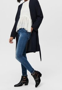 Sorte ankelstøvler med udsnitte stropdesign og sølvhardware, parret med blå skinny jeans og en hvid broderet peplumtop.