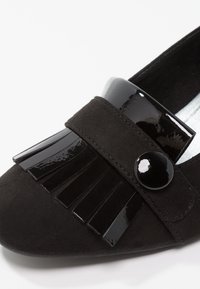 Mocassin en daim noir avec bride en cuir verni noir brillant, détails à franges et un bouton rond brillant sur le dessus avant.