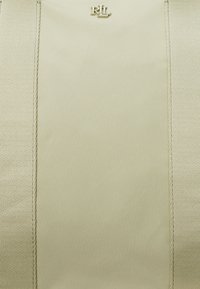 Beige táska sima anyagból, a középső felső részen látható logóval. Kontrasztos varrt szélekkel és texturált oldalsó panelekével rendelkezik.