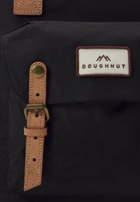 Sac à dos en tissu noir avec une sangle en cuir marron et une boucle en laiton, comportant une étiquette rectangulaire affichant "DOUGHNUT" et un design de montagne.