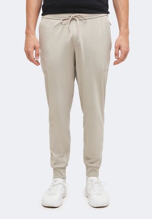 Personne portant un pantalon de jogging beige avec taille à cordon et des baskets blanches, debout devant un fond clair uni.