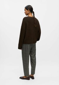 Pull oversized en maille texturée marron, pantalon à carreaux gris, chaussures plates foncées ; mannequin vu de dos avec les cheveux coiffés en tresse.