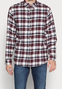 Man som bär en långärmad skjorta med knäppning i ett maroon-, vitt- och svart rutmönster, tillsammans med blå jeans.