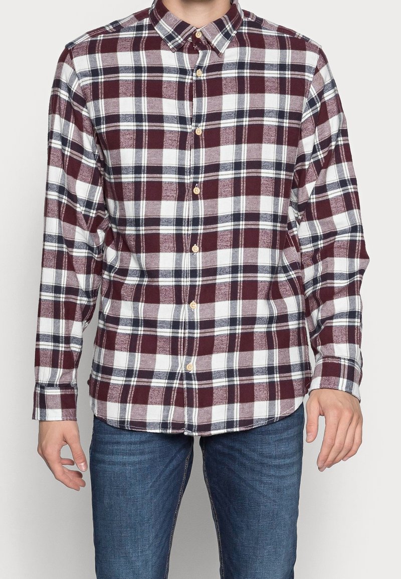 Man som bär en långärmad skjorta med knäppning i ett maroon-, vitt- och svart rutmönster, tillsammans med blå jeans.