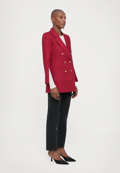 Red double-breasted blazer with gold buttons, side slits, and notched lapels, paired with black straight-leg jeans and black heels.