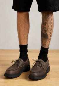 Brown leather shoes with round toes, black rubber soles, and laces. Worn with black socks and shorts, featuring a leg tattoo in the background.