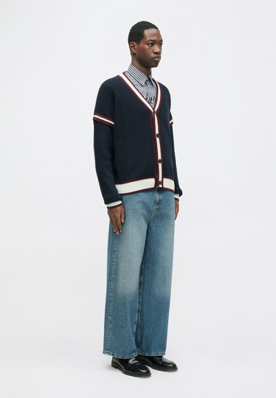 Cardigã de malha azul-marinho com listras vermelhas e brancas, abotoado, por cima de uma camisa listrada. Combinado com jeans largos azul claro e sapatos pretos.