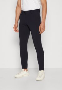 Pantaloni slim-fit blu navy in un tessuto morbido con chiusura a bottone, tasche laterali e un design leggermente aderente, abbinati a sneakers bianche.