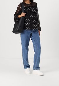 Blouse noire transparente à pois métalliques dorés, associée à un jean en denim bleu ample et des baskets blanches. Accompagnée d'un sac à main noir.