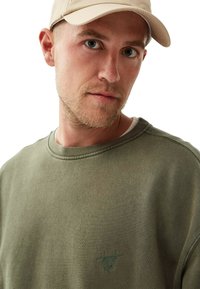 Ung man med ljus skägg som bär en beige keps och en olivgrön sweatshirt med "Ärlighet Härskar" broderat på vit bakgrund.