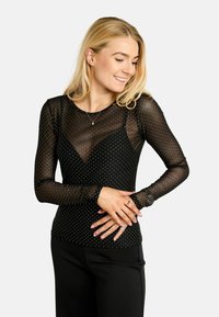 Čierny sieťovaný top s dlhým rukávom a bielymi bodkami, s priliehavým strihom a výstrihom do tvaru V. Nosí sa cez čierne nohavice, čím vytvára vrstvený vzhľad.