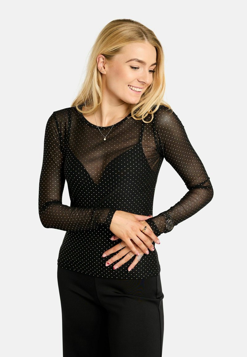 Čierny sieťovaný top s dlhým rukávom a bielymi bodkami, s priliehavým strihom a výstrihom do tvaru V. Nosí sa cez čierne nohavice, čím vytvára vrstvený vzhľad.