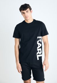 Svart bomullst-shirt med en vit vertikal "KARL"-logotyp på vänstra sidan. Modellen bär den tillsammans med uppvikta svarta shorts.