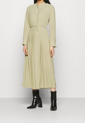 Femme portant une robe midi beige plissée à manches longues avec une ceinture à la taille, associée à des bottines noires à talons brillantes, debout contre un mur uni.