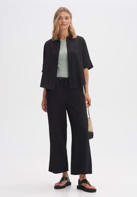 Black textured short-sleeve shirt paired with a light green top, black wide-leg pants, and black sandals. Accessory: woven handbag.
