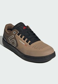 Ruskea ja musta urheilujalkine, jossa on teksturoitu päällinen, rei'itetty varvasosa ja kumipohja. Varustettu harmaalla ja punaisella brändäyksellä.