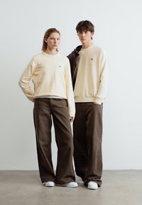 Une femme et un homme se tiennent côte à côte vêtus de pulls crème, de pantalons larges marron et de baskets blanches, sur un fond uni.