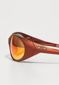 Gafas de sol mate rojas con lentes de color naranja, que presentan un diseño geométrico y un acento con logotipo en la patilla. Forma angular y estilizada.