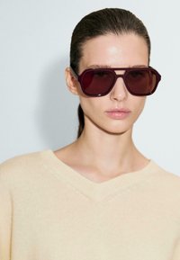Lunettes de soleil avec des verres teintés bordeaux et une forme angulaire, entourées d'une couleur claire contrastante. Portées avec un pull V en jaune pâle.