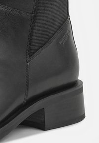 Bota de tornozelo em couro preto com um painel de tecido texturizado, biqueira quadrada e um salto grosso. Apresenta detalhes com o logótipo distintivo na lateral.