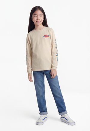 Młoda dziewczyna z długimi ciemnymi włosami, uśmiechająca się, ubrana w beżową bluzę Santa Cruz z niebieskimi grafikami na rękawach, niebieskie dżinsy i białe trampki.