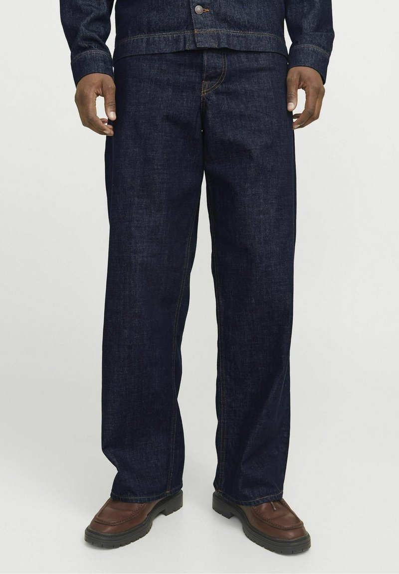 Jean droit bleu foncé porté avec des chaussures en cuir marron et une veste en denim foncé assortie, visible de la moitié du torse vers le bas.