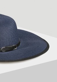 Granatowy słomiany kapelusz z szerokim rondem, teksturowany wzór i szczegóły z czarnej skóry. Posiada okrągła koronę i miękkie wykończenie materiału.