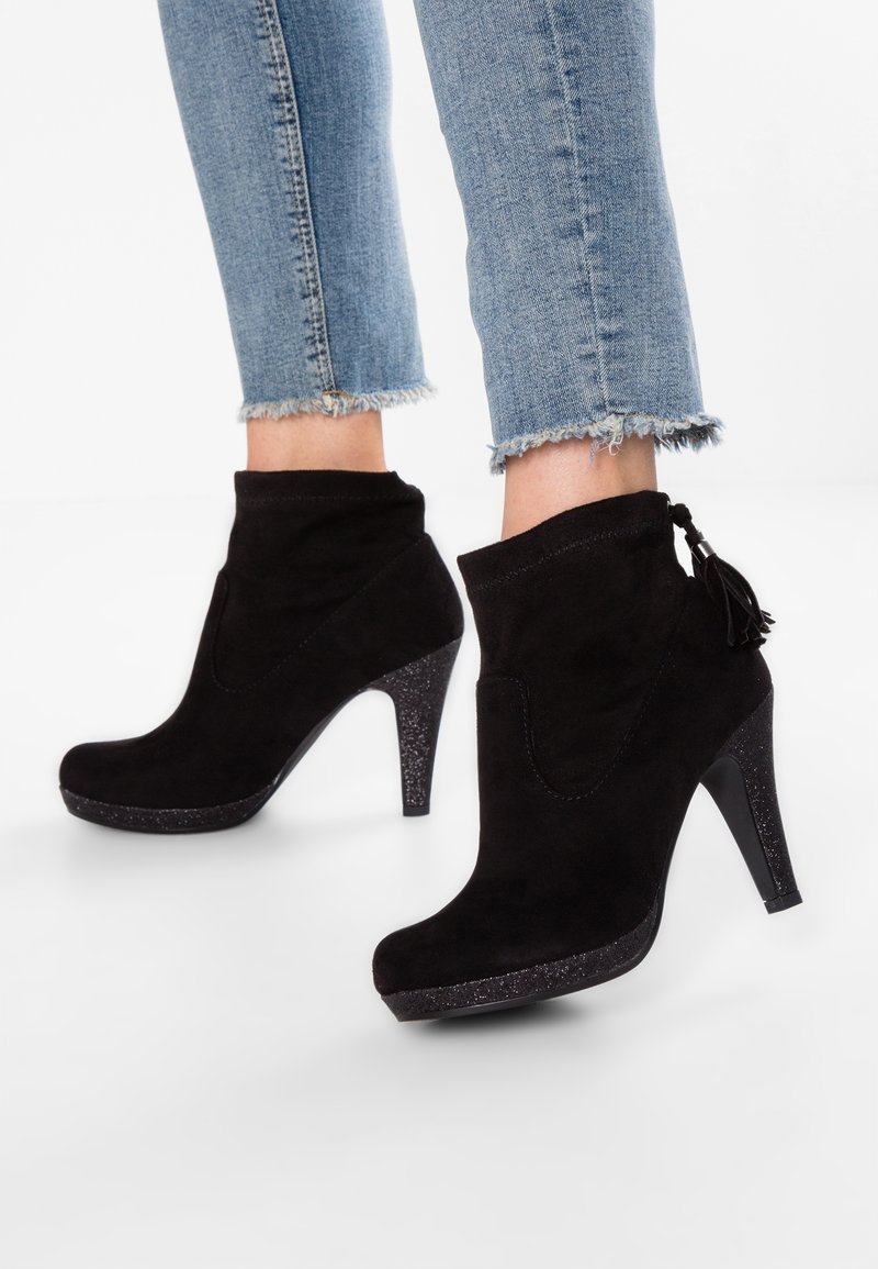 Bottines à talons hauts en suède noir avec talons scintillants et détails à pompons, portées avec un jean bleu à ourlet effiloché.