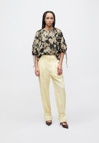 Bluza s cvjetnim uzorkom u crnoj i bež boji s naboranim rukavima i vezicama, uparena s visokim žutim hlačama i smeđim potpeticama.